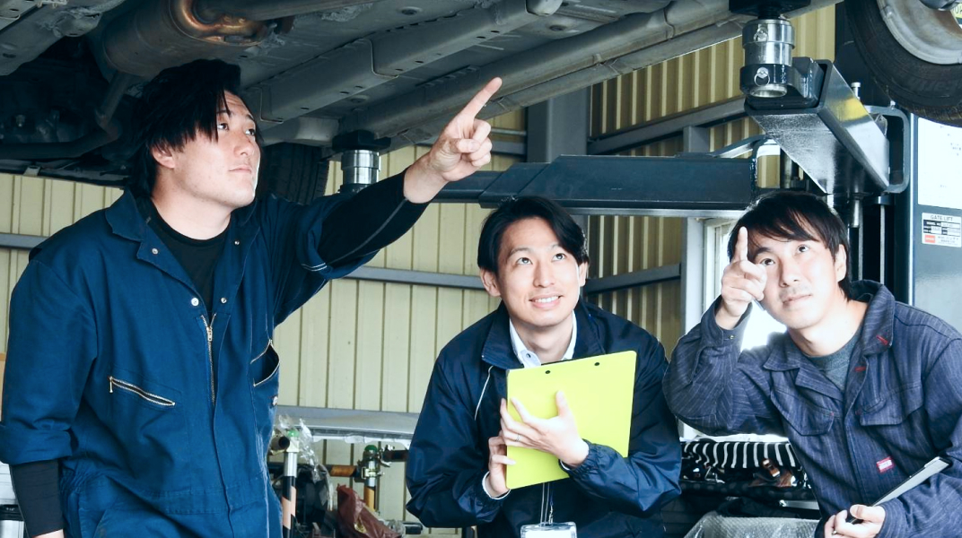 作業服を着た3人の男性がリフトアップされた車の下で指を指しながら点検箇所について話し合っている様子。1人はクリップボードを持っている。