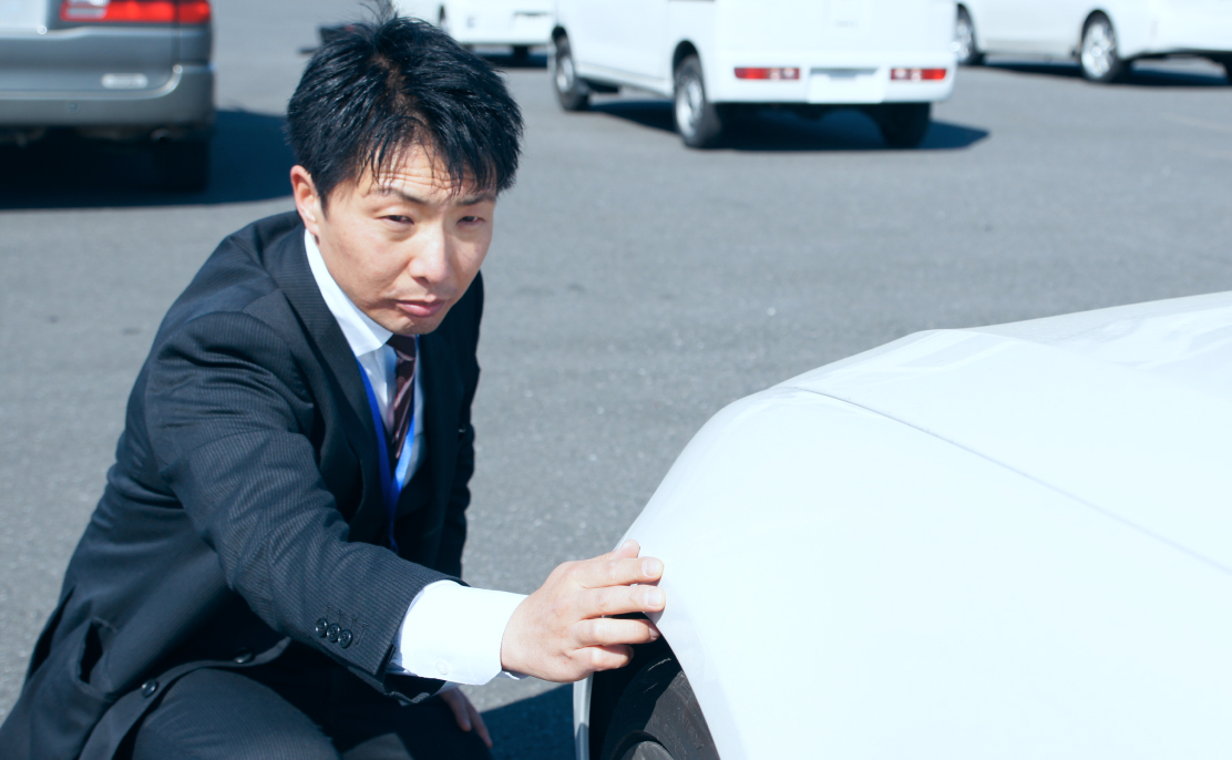 スーツ姿の男性が屋外で車のボディを手で触れながら状態を確認している様子。