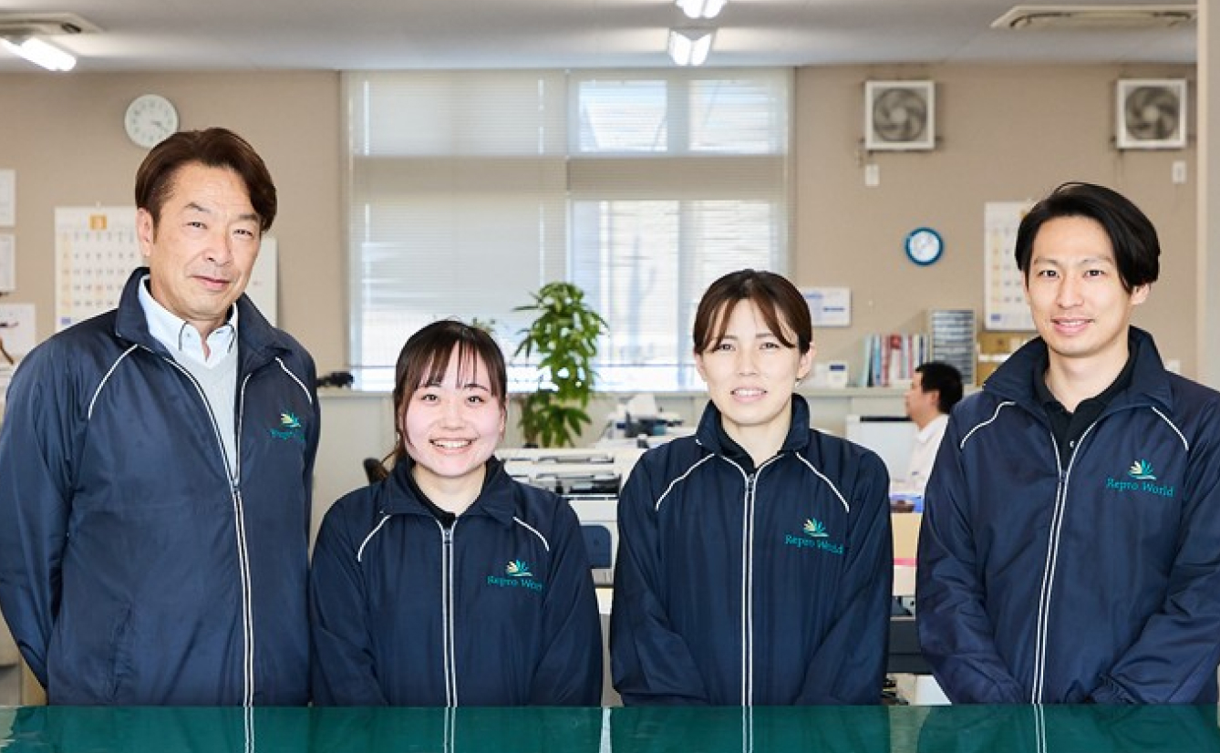 オフィス内で制服を着た4人のスタッフがカメラに向かって笑顔で立っている集合写真。