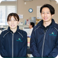 オフィス内で制服を着用した男性と女性が笑顔で並んでいる様子。背景にはデスクや書類棚が写っている。
